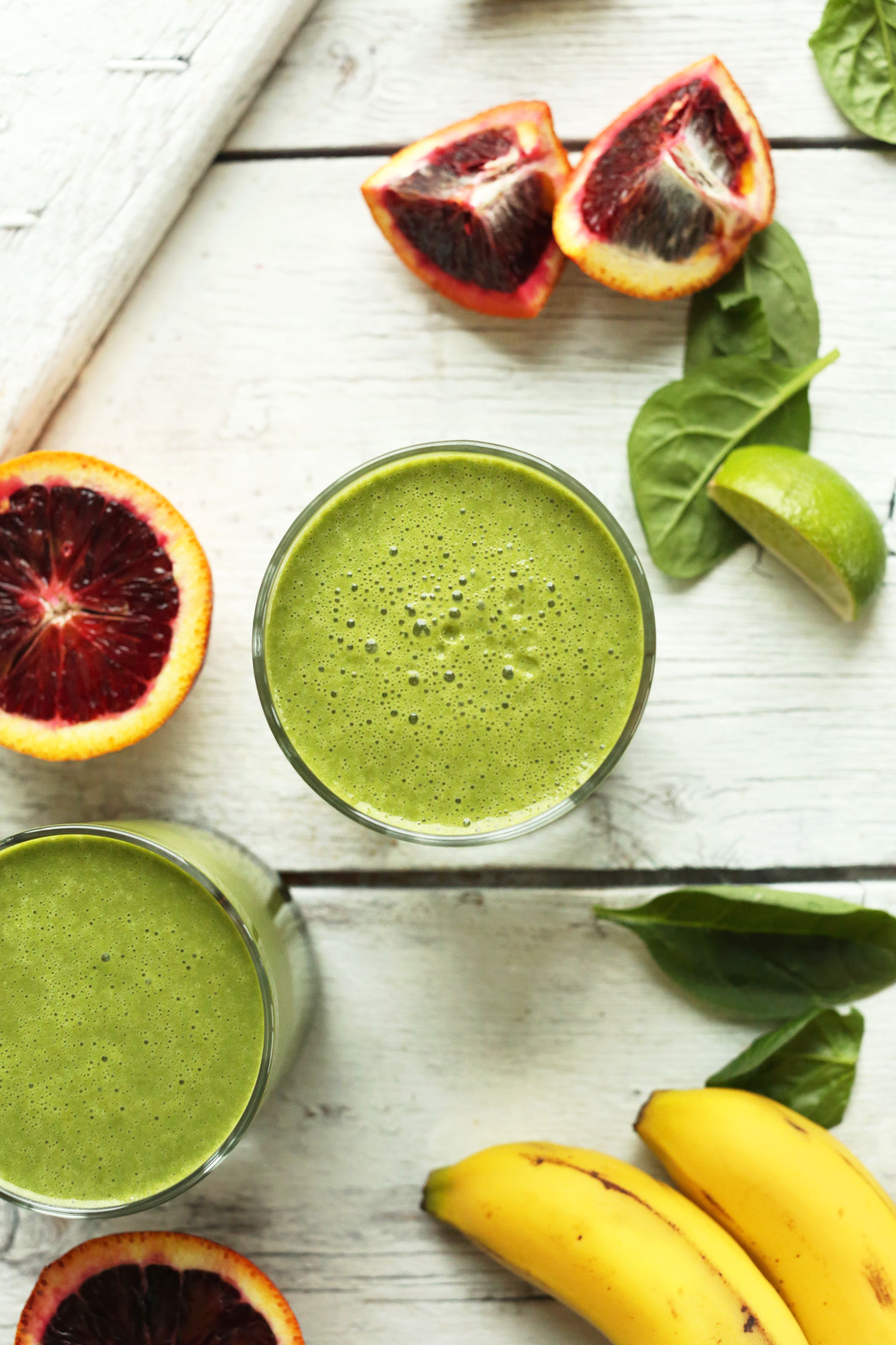 Glasses of our vegan Blood Orange Green Smoothie surrounded by key ingredients such as banana, blood orange, and spinach